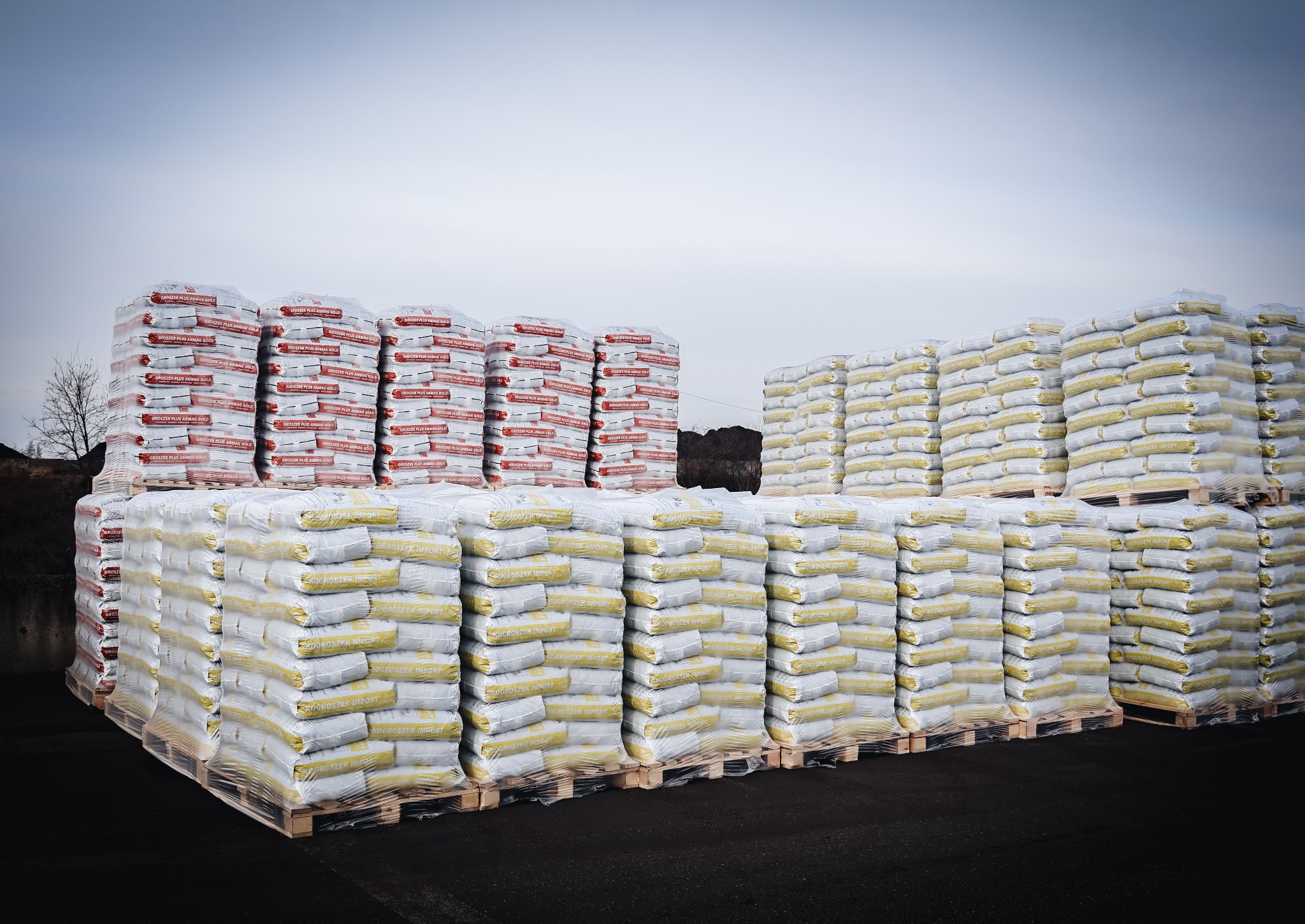 Sztaplowane palety z workami opału na zewnątrz, część z czerwonymi napisami, część z żółtymi, zapakowane w folię, na czarnym asfalcie i pod pochmurnym niebem.