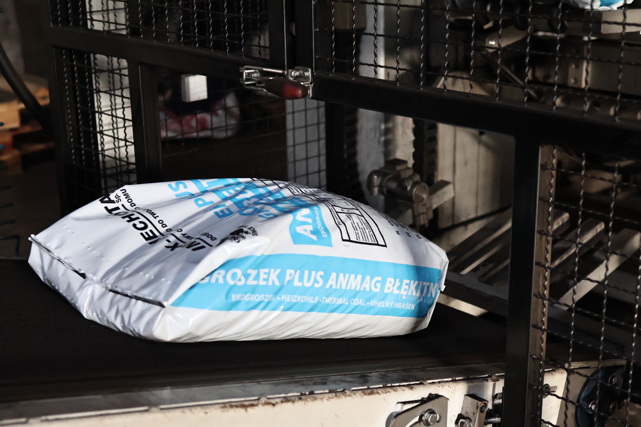 Worek z napisem 'PROSZEK PLUS ANMAG BŁĘKITN' na taśmie produkcyjnej, widoczne elementy maszyny pakującej.