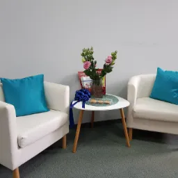 Jasne fotele z turkusowymi poduszkami w poczekalni, stolik z kwiatami i ciasteczkami, ulotki w tle.