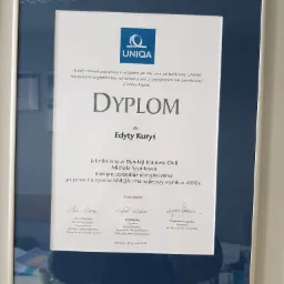 Oprawiony dyplom firmy UNIQA dla Edyty Kuryś za najlepszą sprzedaż ubezpieczeń grupowych na życie w 2018 roku, z podpisami gratulacyjnymi, zawieszony na ścianie.