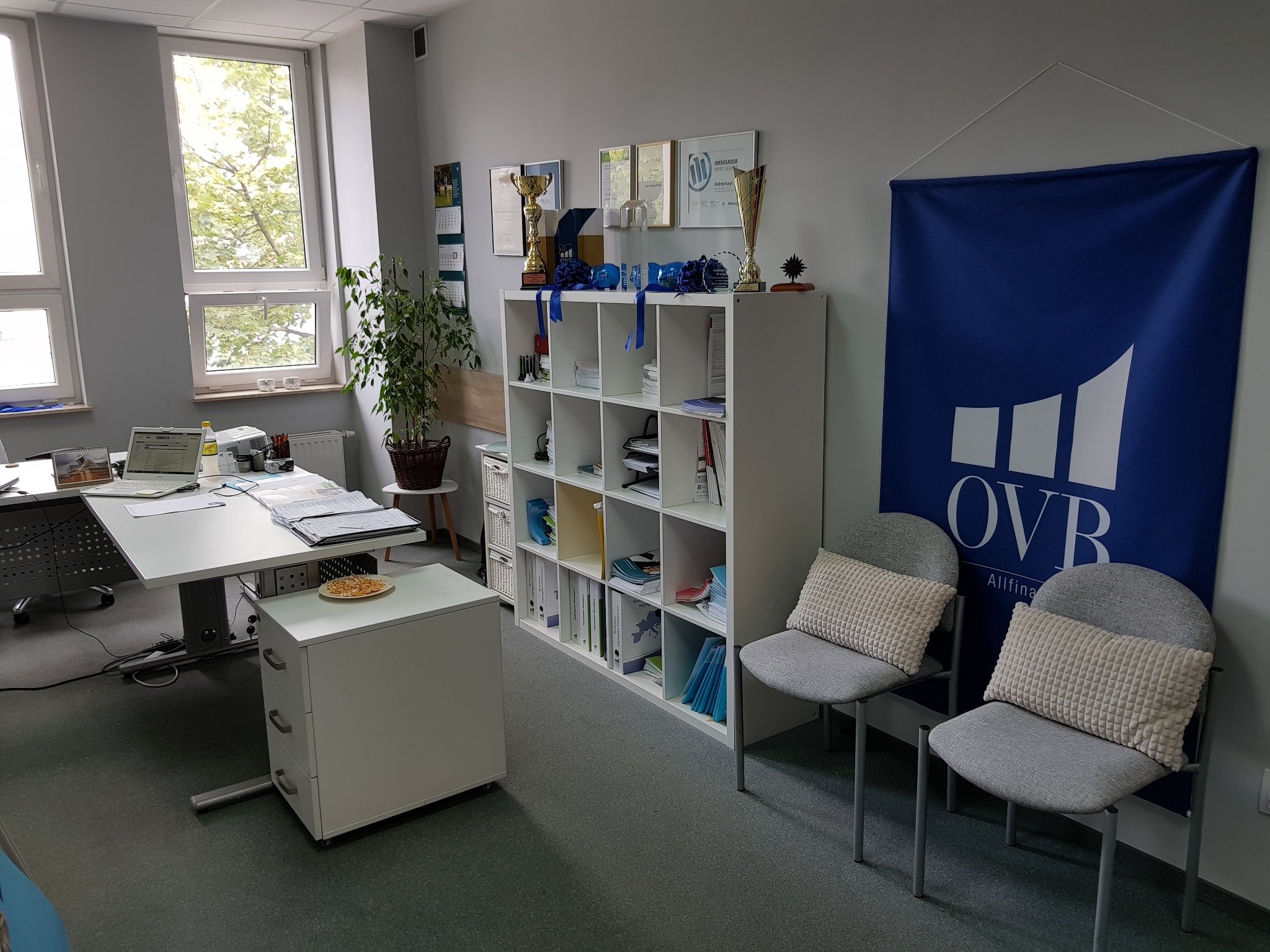Jasne biuro z biurkiem, regałem z trofeami i dyplomami, dwoma krzesłami z poduszkami na tle baneru OVB Allfinanz.