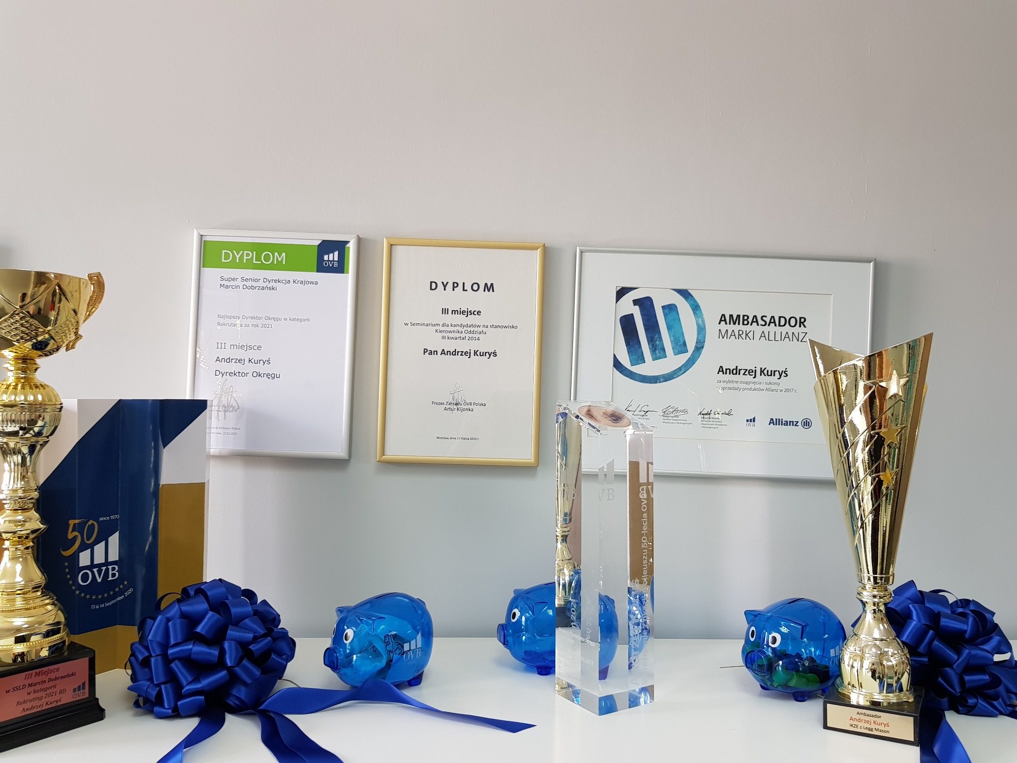 Półka z trofeami, dyplomami i statuetkami firmy OVB i Allianz, wraz z dekoracyjnymi świnkami skarbonkami i kokardami, symbolizującymi sukcesy w rekrutacji i sprzedaży produktów finansowych.