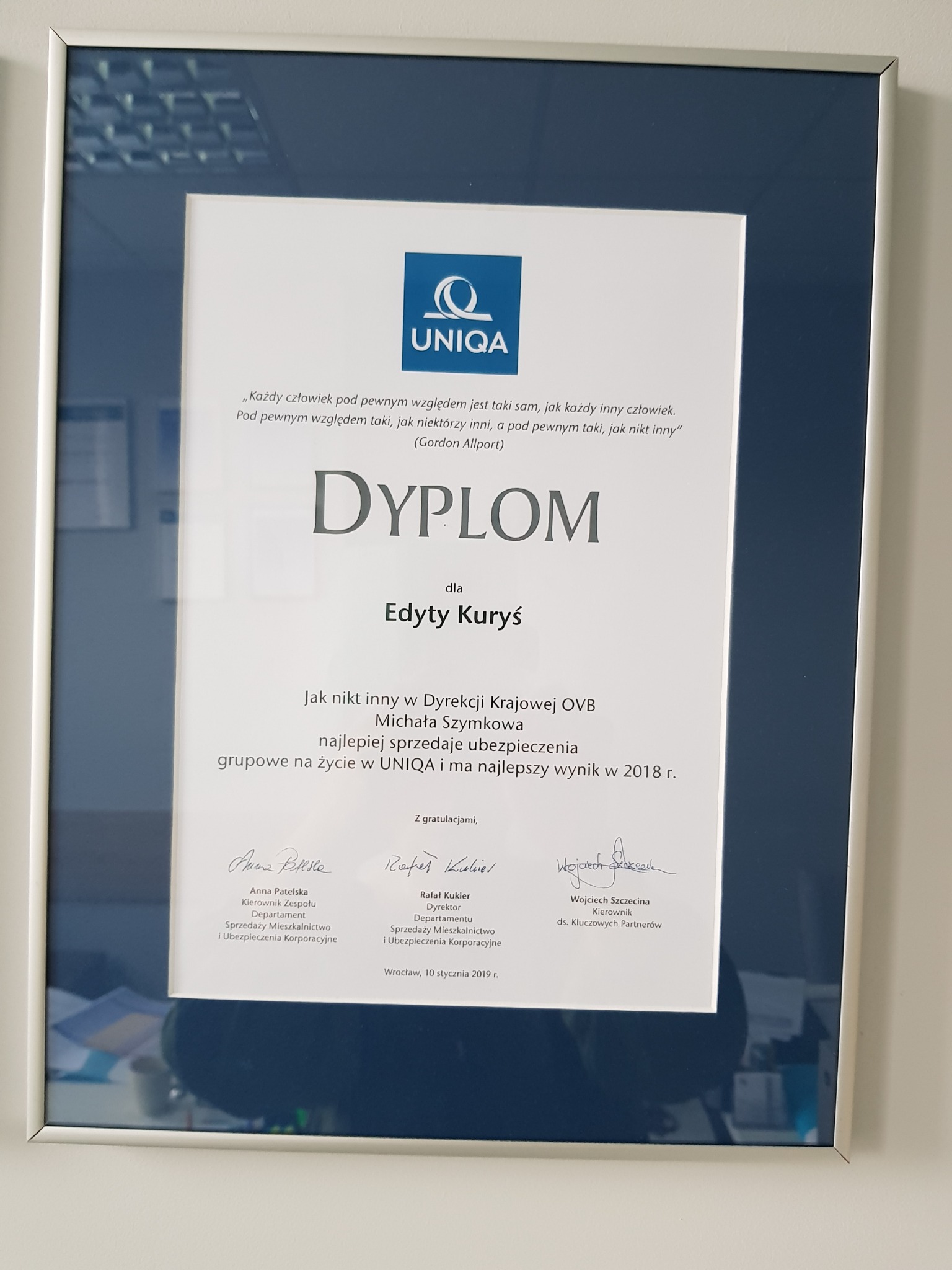 Oprawiony dyplom firmy UNIQA dla Edyty Kuryś za najlepszą sprzedaż ubezpieczeń grupowych na życie w 2018 roku, z podpisami gratulacyjnymi, zawieszony na ścianie.