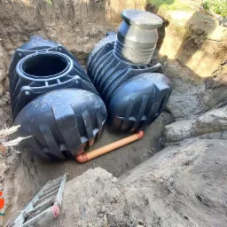 Dwa czarne zbiorniki z tworzywa sztucznego, połączone pomarańczową rurą, umieszczone w wykopie ziemnym, z widoczną drabinką i logo firmy w rogu.