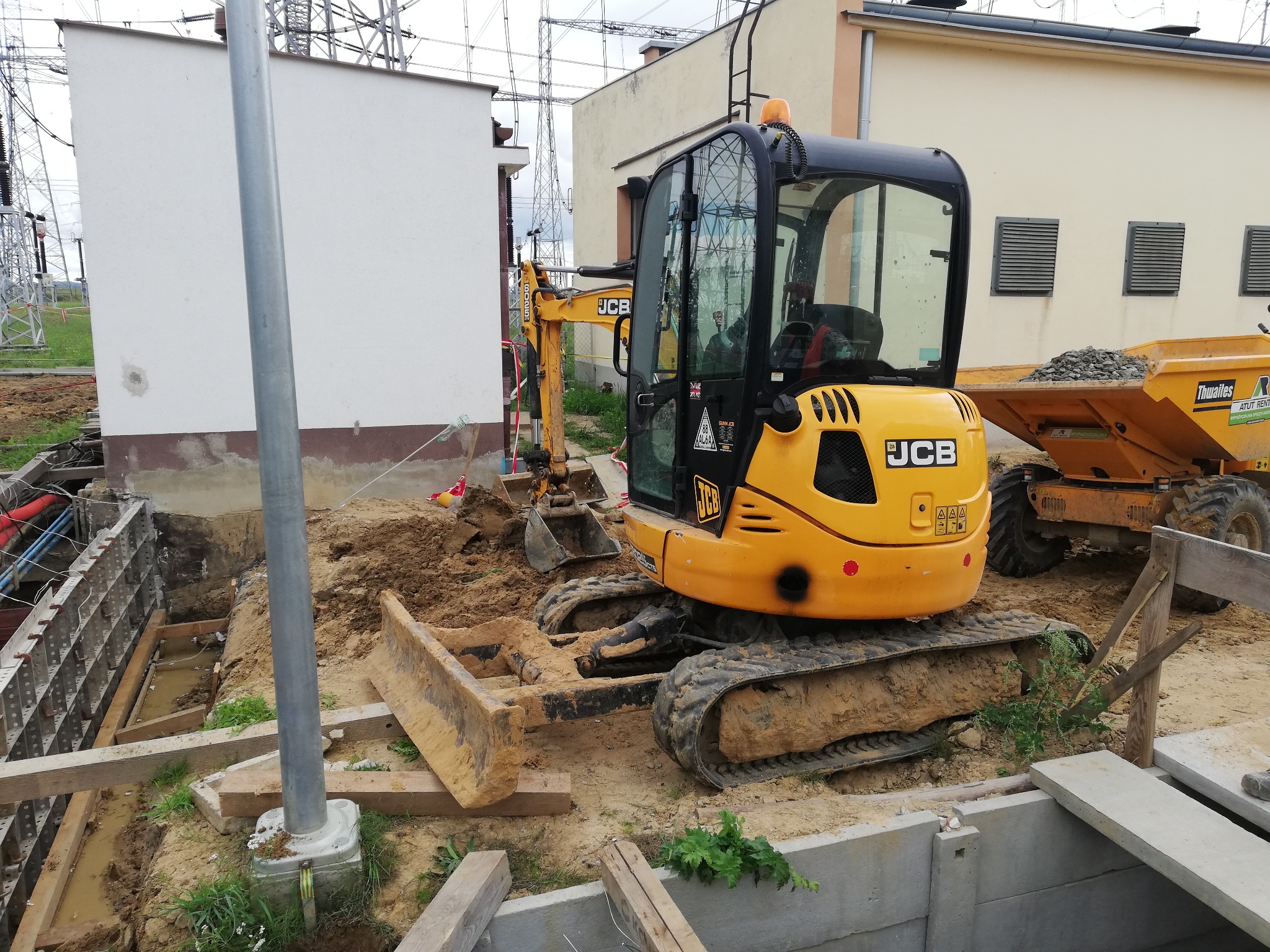 Żółta mini koparka JCB na placu budowy, widoczne wykopy z szalunkiem i ziemia, w tle słupy wysokiego napięcia i budynek.
