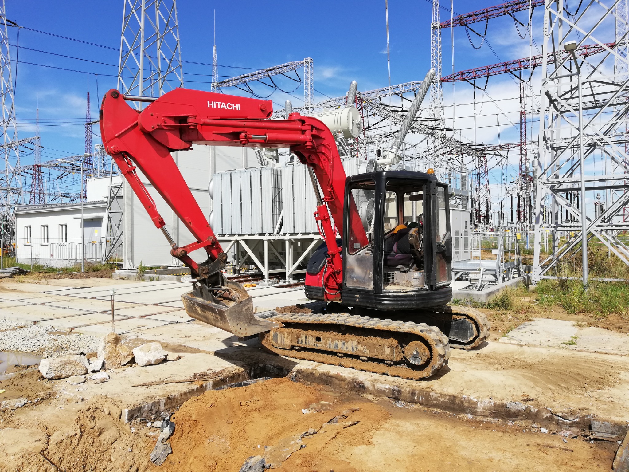 Czerwono-czarna minikoparka Hitachi z operatorem, na placu budowy z wykopem ziemnym, na tle stacji transformatorowej i błękitnego nieba.