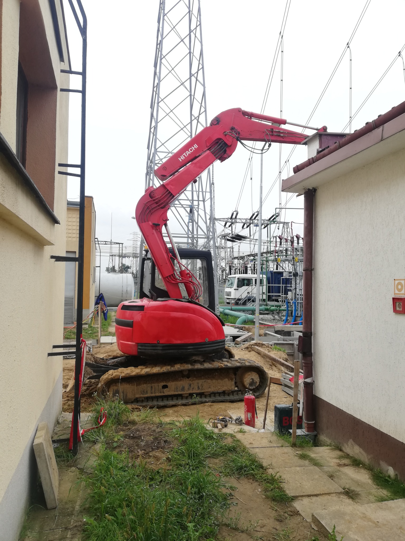 Czerwona koparka Hitachi na placu budowy, ramię skierowane w stronę dachu budynku, w tle stacja transformatorowa i słup wysokiego napięcia.