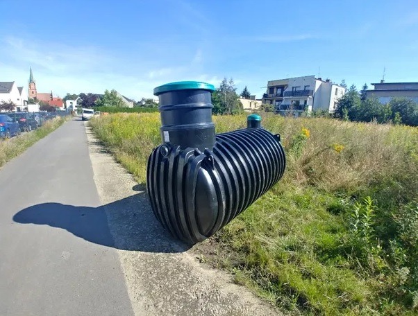 Czarny zbiornik z tworzywa sztucznego, prawdopodobnie na ścieki, leży na skraju drogi asfaltowej, obok zarośniętego terenu z widocznymi budynkami w tle.