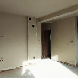 Pokój w trakcie prac wykończeniowych: ściany pokryte świeżym, jasnym tynkiem, widoczne puszki elektryczne i otwory wentylacyjne, podłoga zabezpieczona folią, ślady po tynkowaniu przy podłodze.