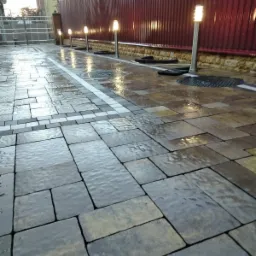 Ułożona kostka brukowa w odcieniach szarości i beżu, mokra od deszczu, z dekoracyjnym pasem jasnoszarej kostki i oświetleniem wzdłuż ogrodzenia z blachy trapezowej.