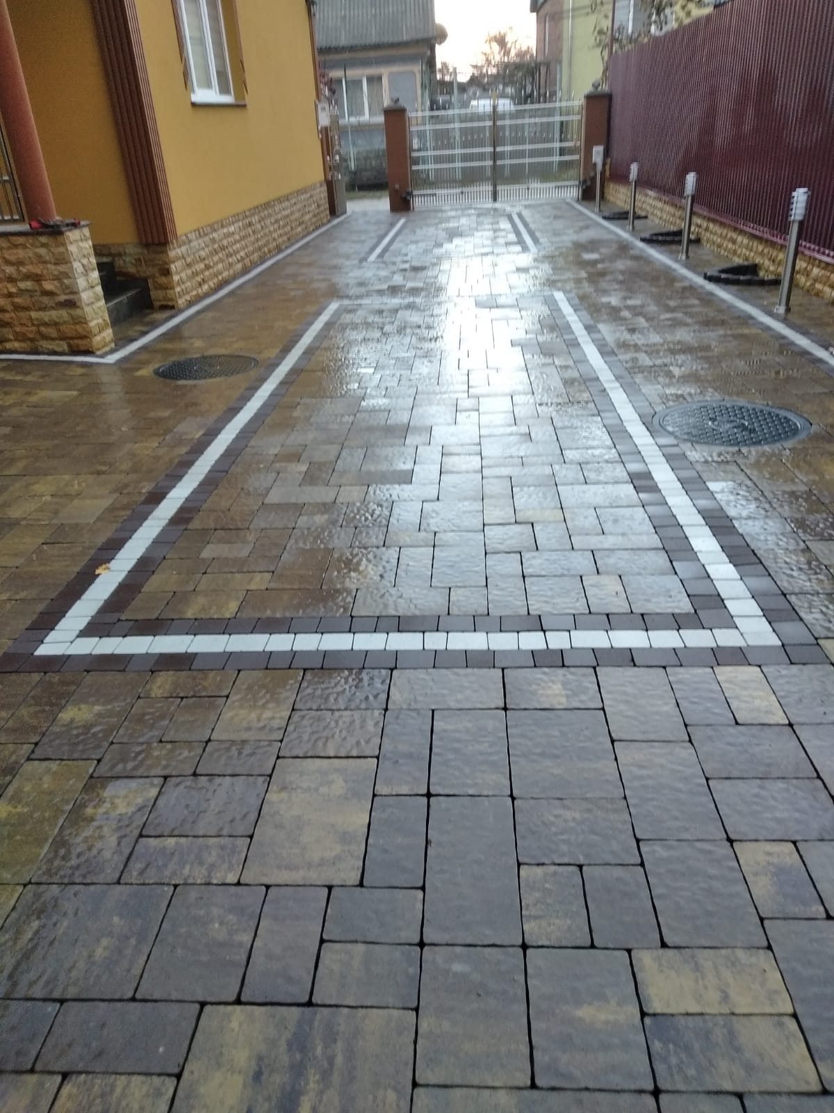 Ułożona kostka brukowa w odcieniach brązu i szarości, obramowana białymi i ciemnobrązowymi kostkami, tworząca wzór na podjeździe przed domem z widoczną studzienką kanalizacyjną i metalową bramą w tle.