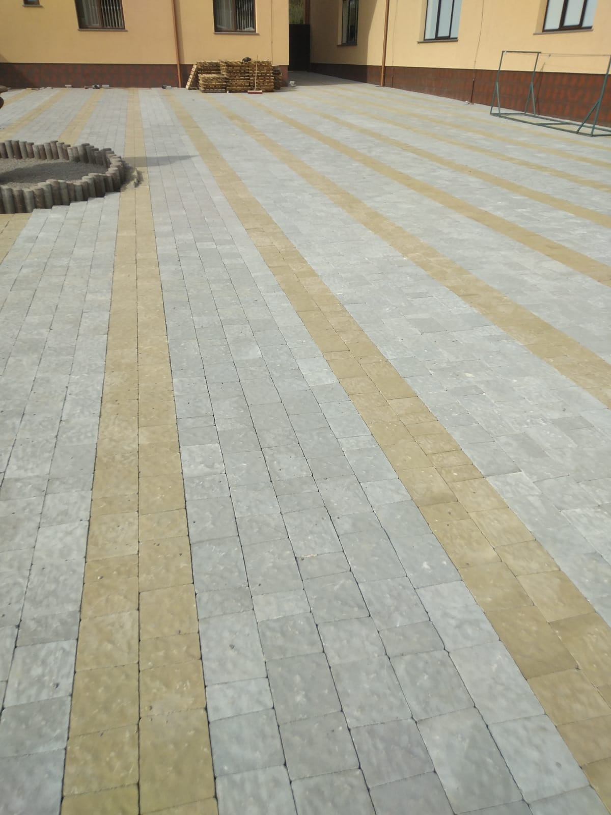 Ułożona kostka brukowa w dwóch odcieniach szarości i beżu, tworząca wzór pasów na podwórku przed budynkiem z paletą kostki w tle.