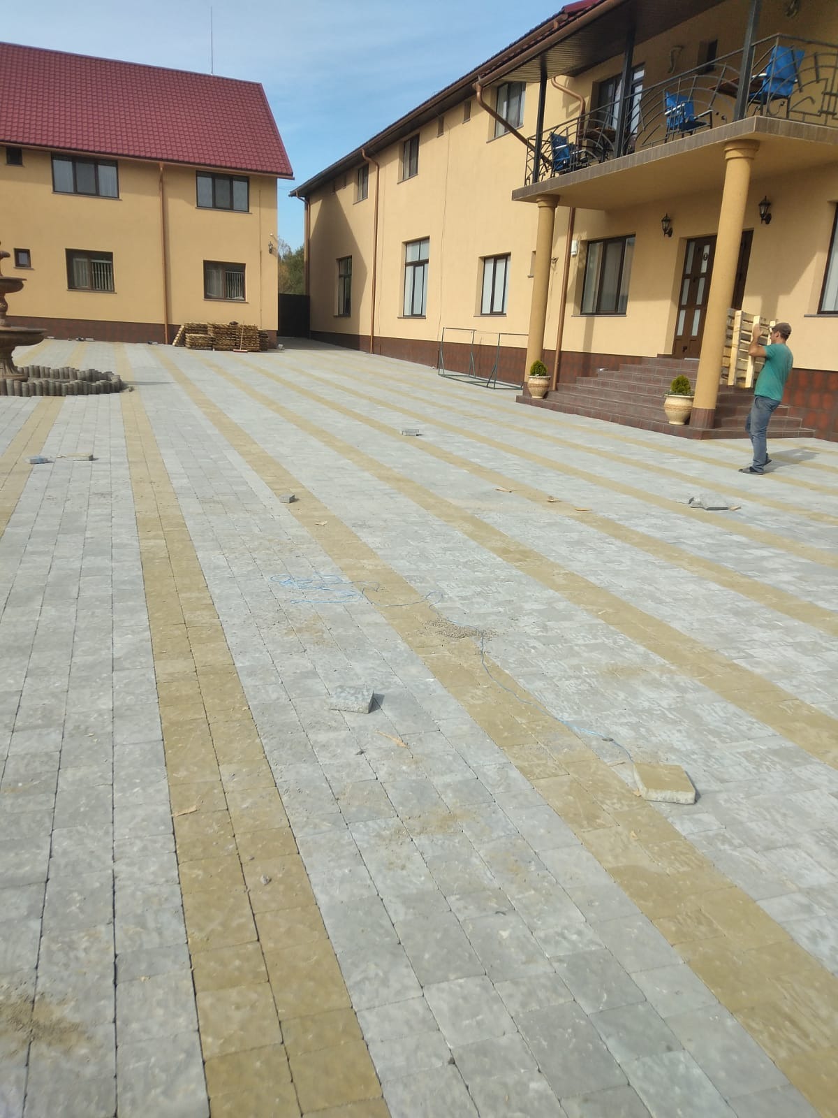 Nowo ułożona kostka brukowa w odcieniach szarości i beżu przed budynkiem z balkonem, z widocznymi niedokończonymi fragmentami i pracownikiem przenoszącym paletę w tle.