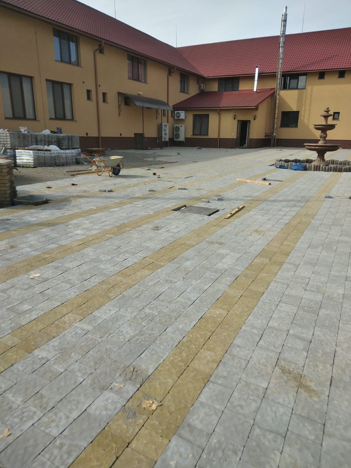 Plac z ułożoną częściowo kostką brukową w szaro-żółte pasy, w tle budynek z czerwoną dachówką, taczka i fontanna.