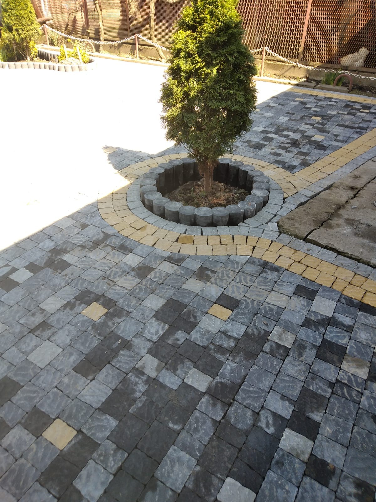 Szaro-czarna kostka brukowa ułożona we wzór, z żółtymi akcentami, otaczająca małe drzewko w okrągłej obudowie z betonowych elementów.