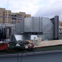 Better Climate Sp. z o.o. - Montaż dużych, srebrnych kanałów wentylacyjnych na dachu budynku mieszkalnego, widoczne narzędzia i materiały izolacyjne.