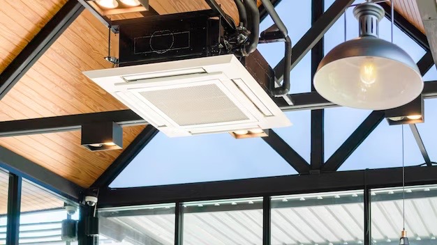 Kasetonowa jednostka klimatyzacyjna zamontowana na drewnianym suficie z widoczną konstrukcją stalową i oświetleniem punktowym w nowoczesnym pomieszczeniu z dużymi oknami.