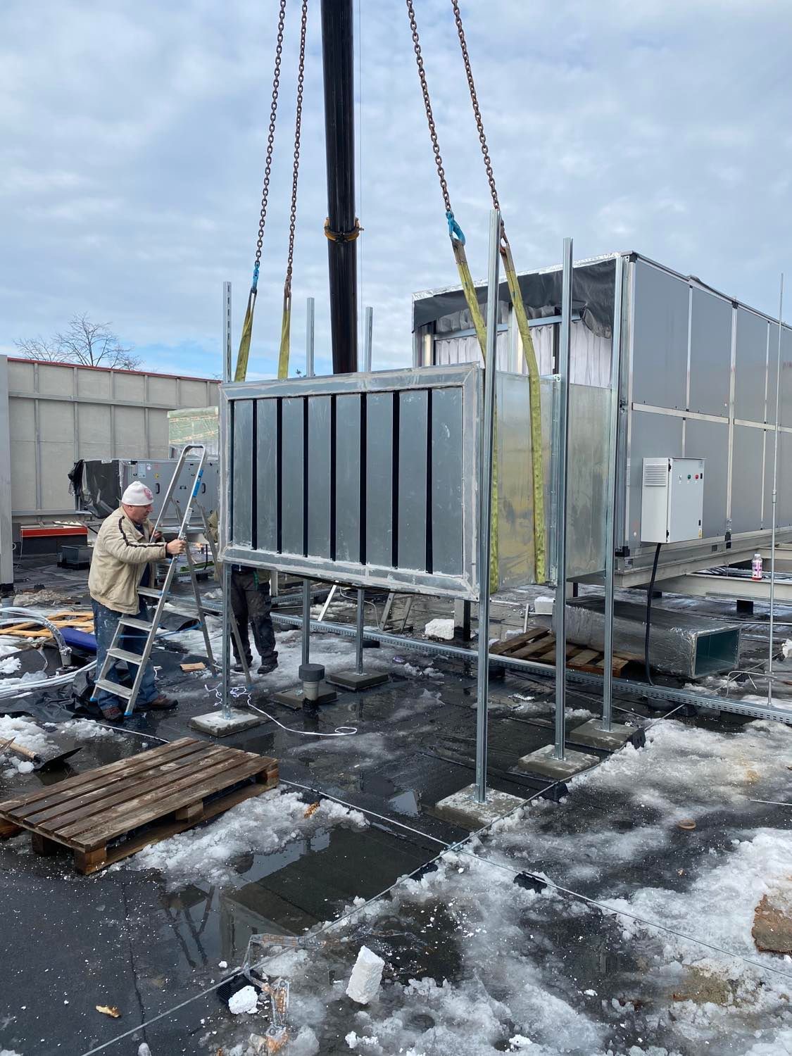 Montaż centrali wentylacyjnej na dachu budynku z użyciem dźwigu, widoczny pracownik na drabinie i pozostałości śniegu.