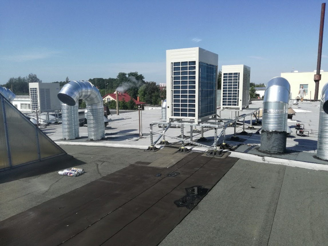 Instalacja wentylacyjna i klimatyzacyjna na płaskim dachu budynku komercyjnego, widoczne elementy rekuperacji i klimatyzacji, w tle komin z dymem.