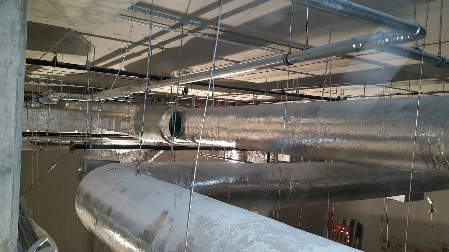 Widok z dołu na system rur wentylacyjnych i klimatyzacyjnych pod sufitem, z widocznym metalowym stelażem podtrzymującym i izolacją termiczną na rurach.