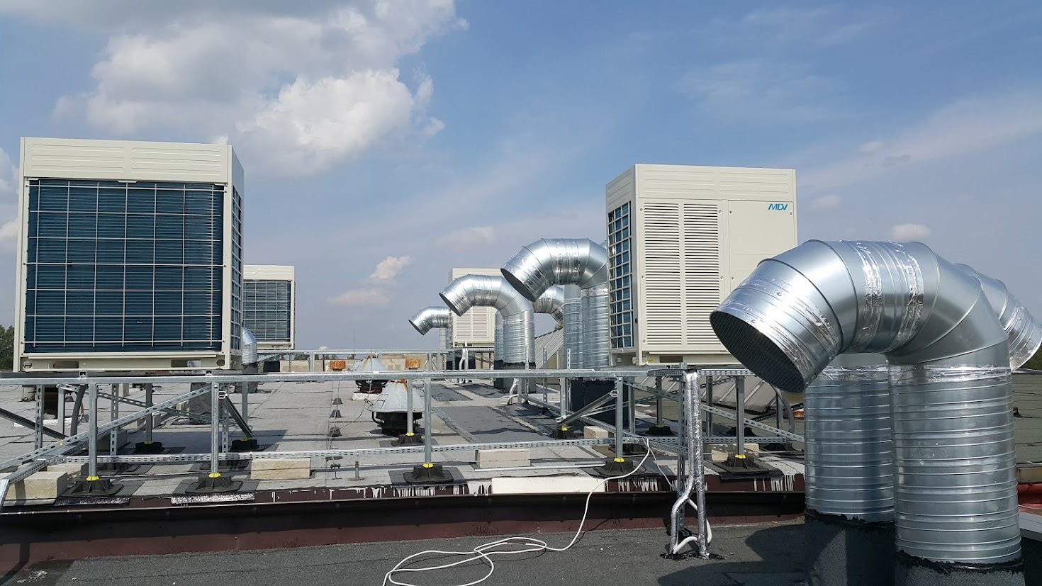 Instalacja klimatyzacji na dachu budynku komercyjnego z widocznymi jednostkami zewnętrznymi i kanałami wentylacyjnymi, zabezpieczona metalową konstrukcją, widok w słoneczny dzień.