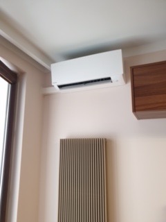 Biały klimatyzator ścienny zamontowany w rogu pokoju, widoczny grzejnik panelowy i fragment okna.