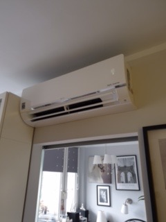 Biały klimatyzator ścienny zamontowany nad przejściem, widok z dołu, w tle okno i dekoracje ścienne.