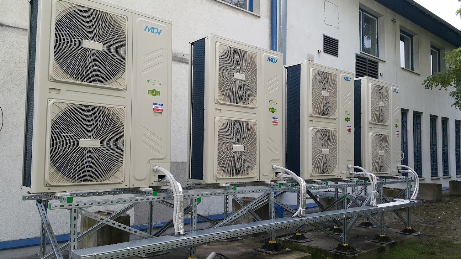Rząd zewnętrznych jednostek klimatyzacyjnych MDI, zamontowanych na metalowej konstrukcji wsporczej przy budynku z oknami.