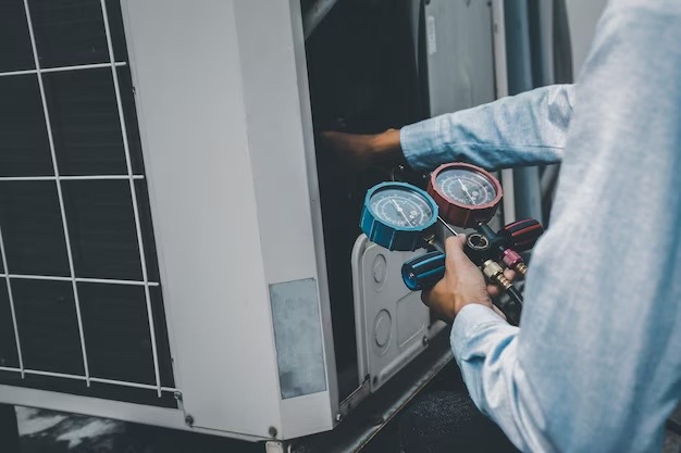 Mężczyzna w jasnej koszuli sprawdza ciśnienie w agregacie chłodniczym za pomocą manometru z dwoma zegarami, niebieskim i czerwonym, podczas serwisu urządzenia.