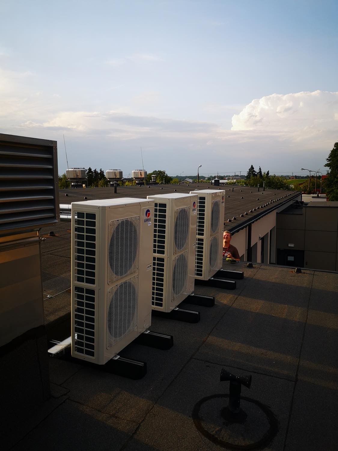 Rząd trzech klimatyzatorów Gree ustawionych na dachu budynku z widocznym fragmentem twarzy instalatora w tle i pochmurnym niebem.