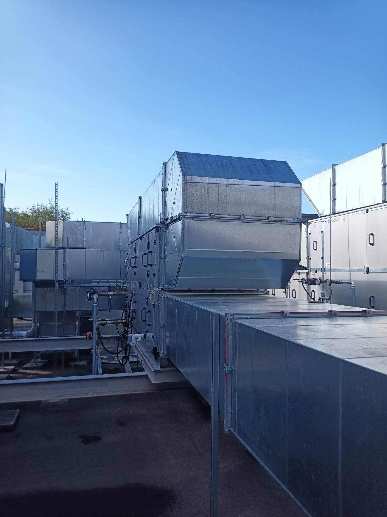 Złożony system kanałów wentylacyjnych i central klimatyzacyjnych na dachu budynku, widok z dołu, niebieskie niebo w tle.