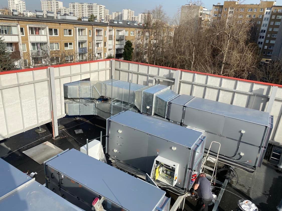 Instalacja przemysłowej wentylacji na dachu budynku, widoczne kanały wentylacyjne, urządzenia i pracownik serwisu przy otwartej skrzynce sterowniczej, w tle zabudowa miejska.