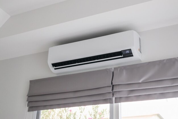 Biały klimatyzator ścienny z wyświetlaczem temperatury 28 stopni Celsjusza, zamontowany nad szarymi roletami rzymskimi w jasnym pomieszczeniu.