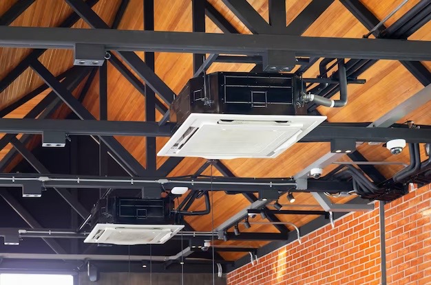 Kasetonowe klimatyzatory sufitowe zamontowane na czarnej, stalowej konstrukcji pod drewnianym stropem w pomieszczeniu z fragmentem ceglanej ściany.
