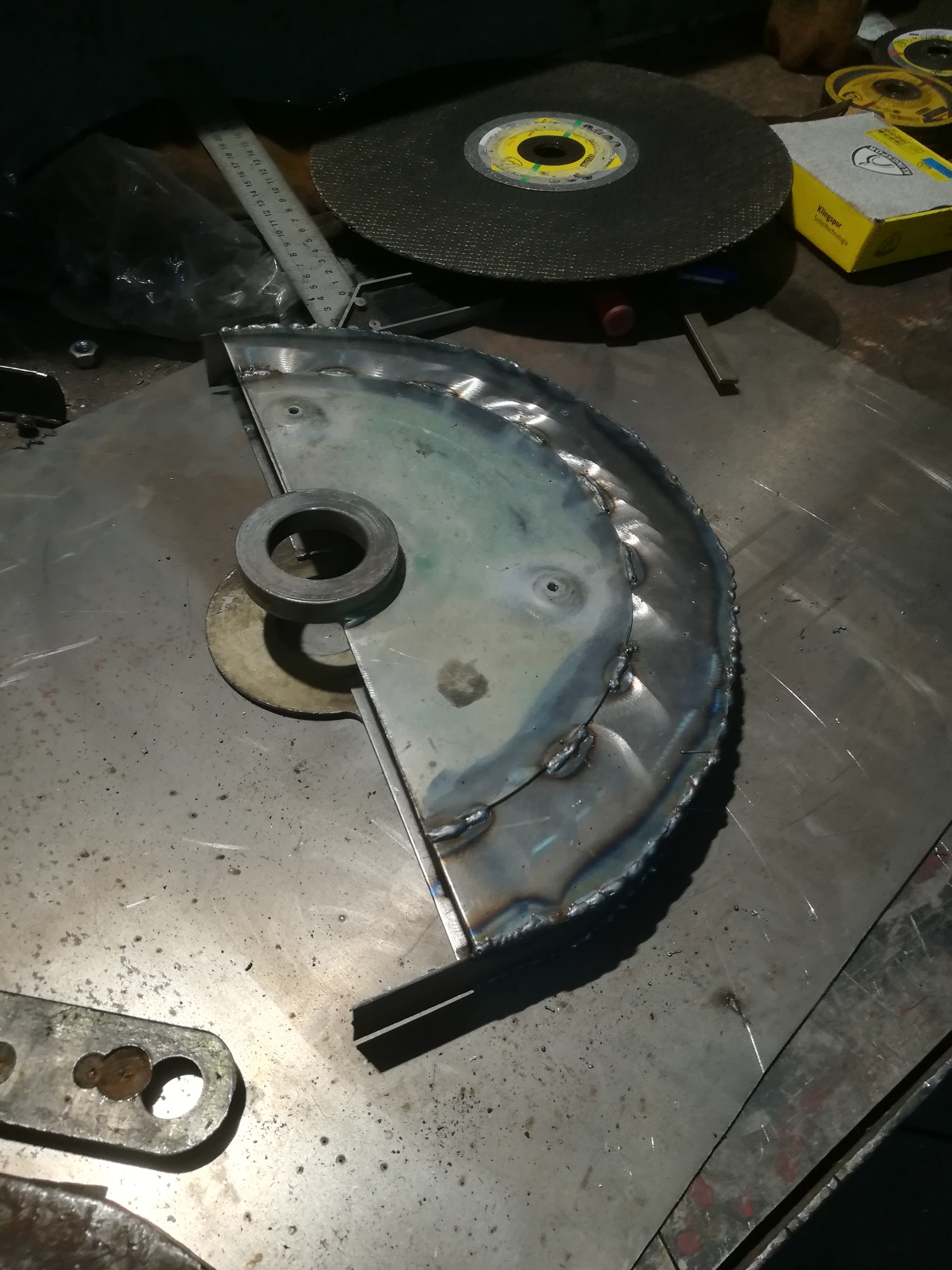 Półokrągła metalowa osłona tarczy szlifierskiej, spawana punktowo, leżąca na metalowym stole warsztatowym, obok tarcza szlifierska i pudełko po tarczach szlifierskich.