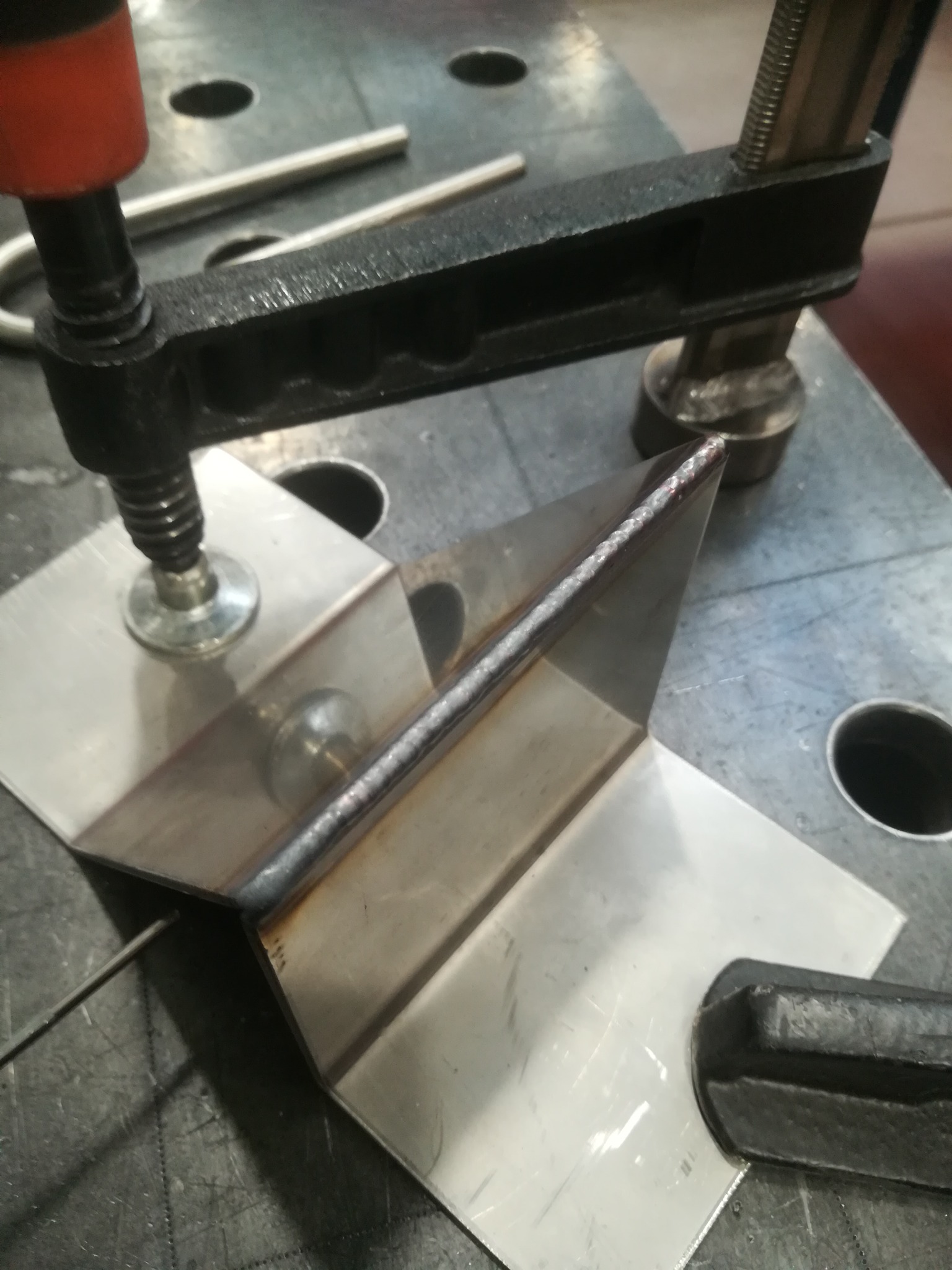 Metalowe elementy zespawane pod kątem prostym, zabezpieczone ściskiem stolarskim na perforowanym stole spawalniczym, widoczny świeży spaw.