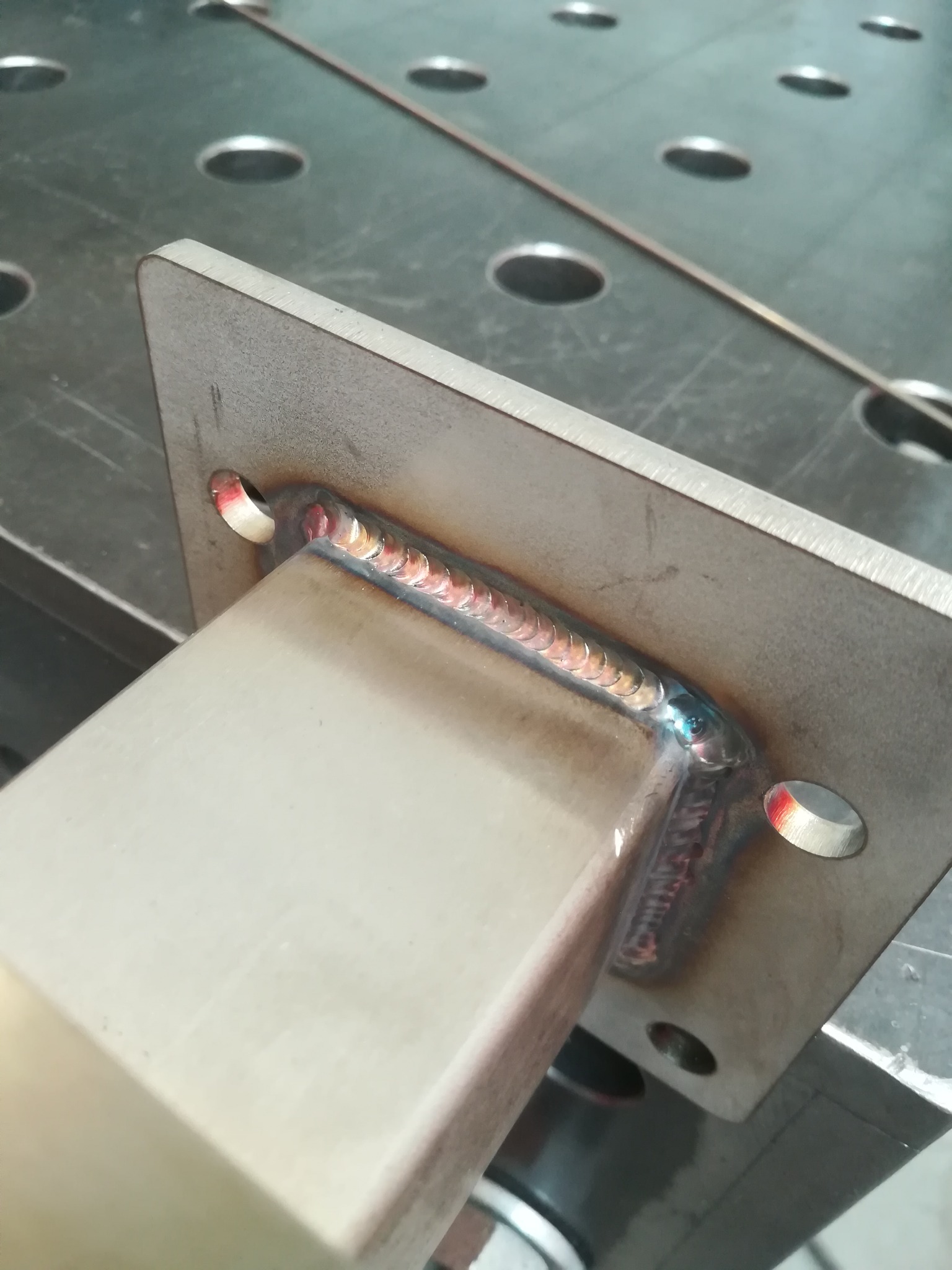 Spoina spawalnicza łącząca kwadratowy profil z płytą z otworami, widoczna na stole spawalniczym z widocznym drutem spawalniczym w tle.