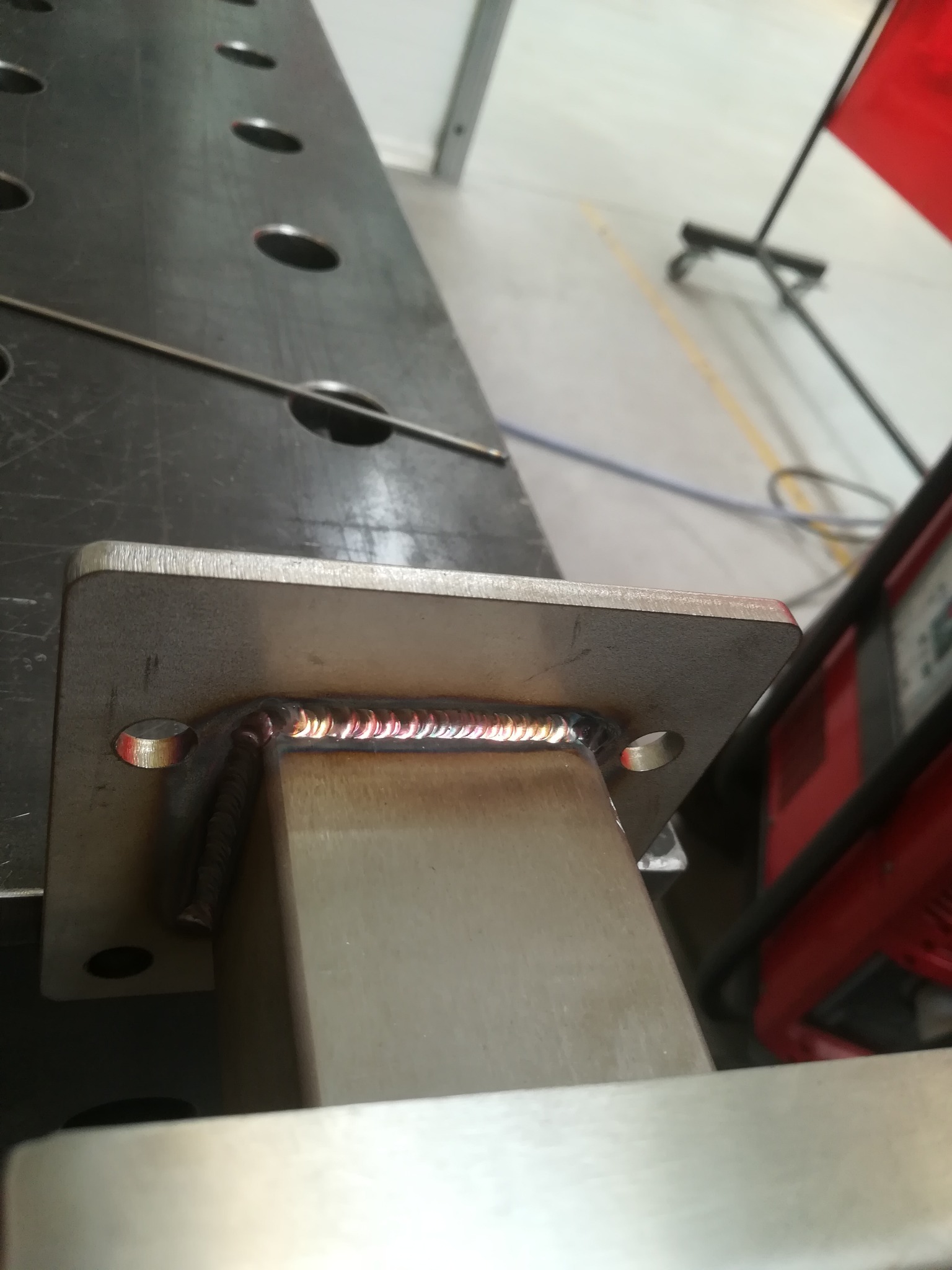 Spoina pachwinowa łącząca stalowy profil kwadratowy z płytą, widoczne regularne fale spoiny, otwory w płycie i narzędzia na stole spawalniczym w tle.