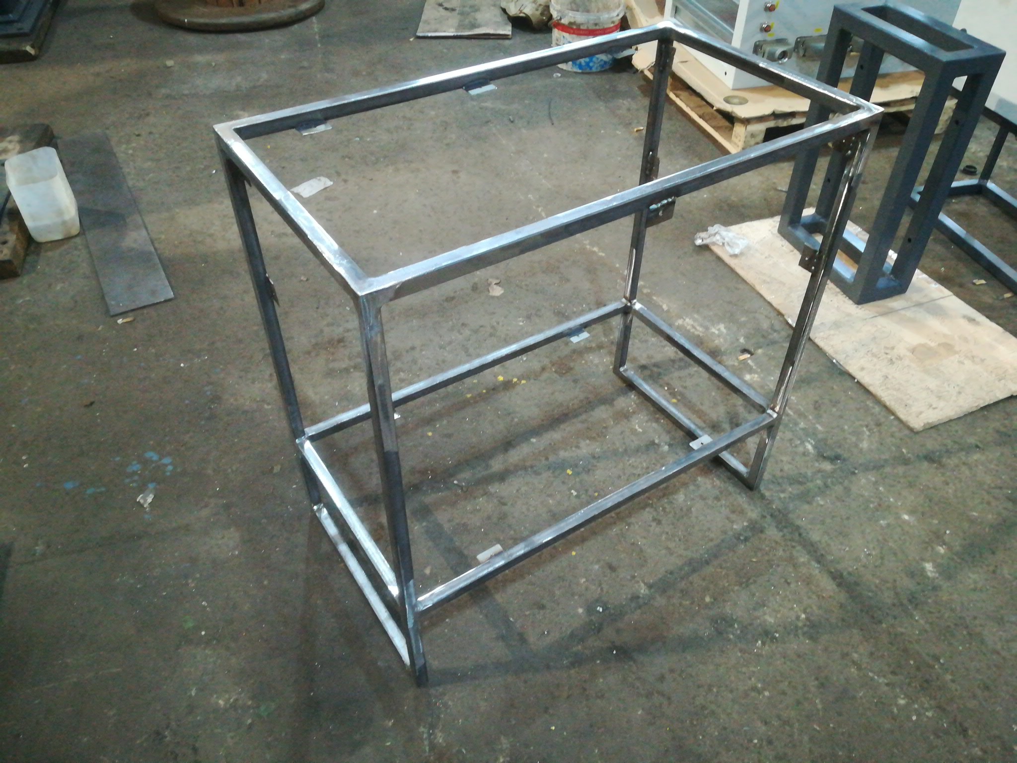 Metalowa rama stołu spawana z profili kwadratowych, widoczne spawy na łączeniach, surowy stan, w tle inne konstrukcje metalowe i drewniana paleta.