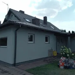 Dom jednorodzinny z szarą elewacją po odświeżeniu, widoczne narzędzia malarskie i drabina przy wejściu, zieleń ogrodu w tle.