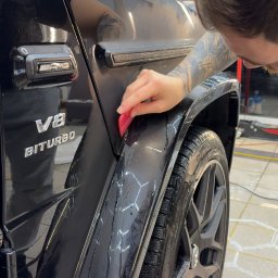 Emka Detailing Studio - Czyszczenie błotnika czarnego Mercedesa G-klasy AMG V8 Biturbo w myjni. Widoczne ślady wody i piany, narzędzie w dłoni osoby z tatuażem na ramieniu.