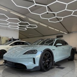 Emka Detailing Studio - Błękitny Porsche Taycan z czarnymi felgami w jasnym pomieszczeniu z heksagonalnym oświetleniem sufitowym. Widoczny fragment innego auta w tle.