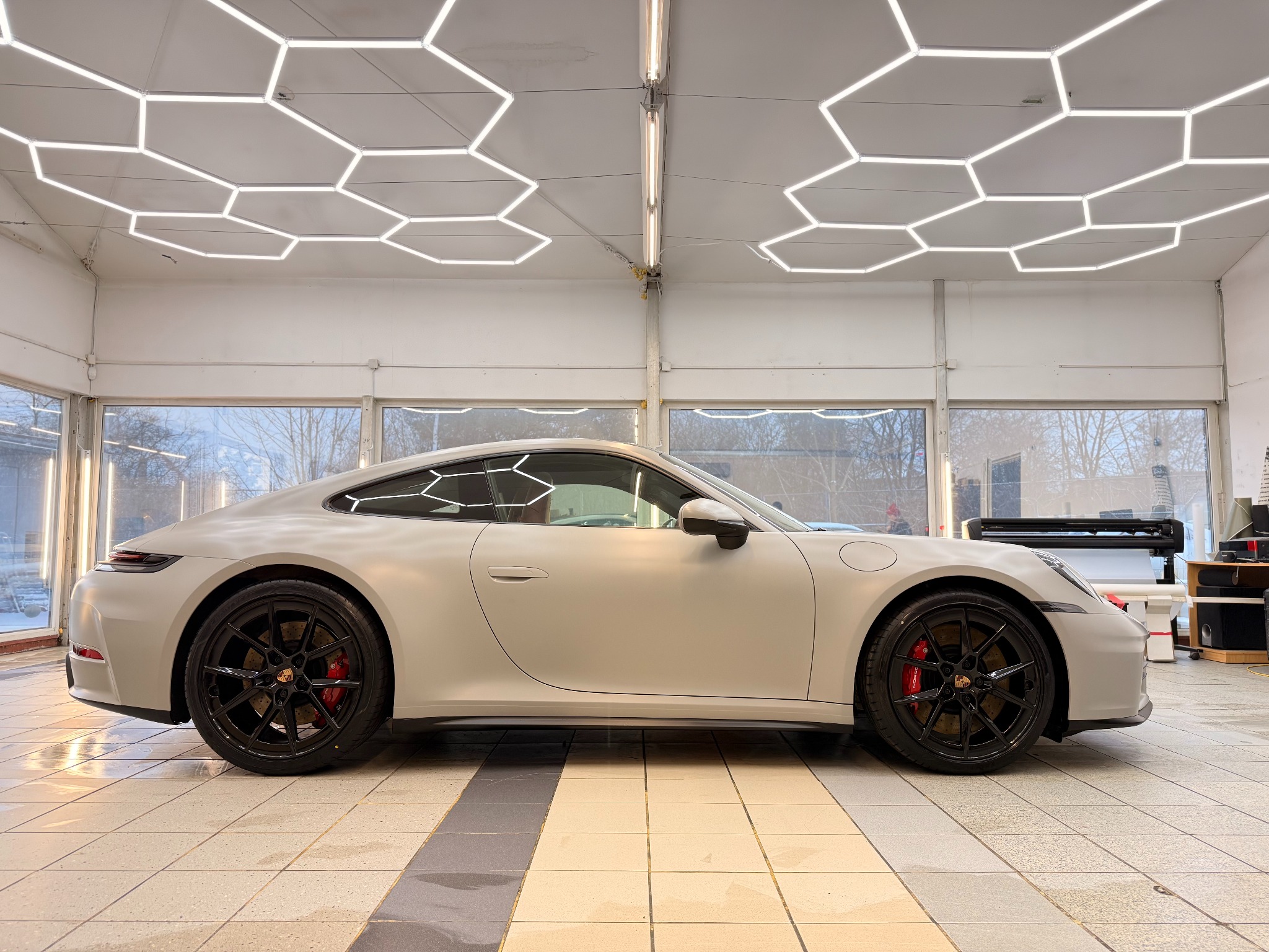 Szary Porsche 911 po oklejeniu folią PPF Hexis, widok z boku w jasnym pomieszczeniu z heksagonalnym oświetleniem sufitowym i czarnymi felgami z czerwonymi zaciskami.