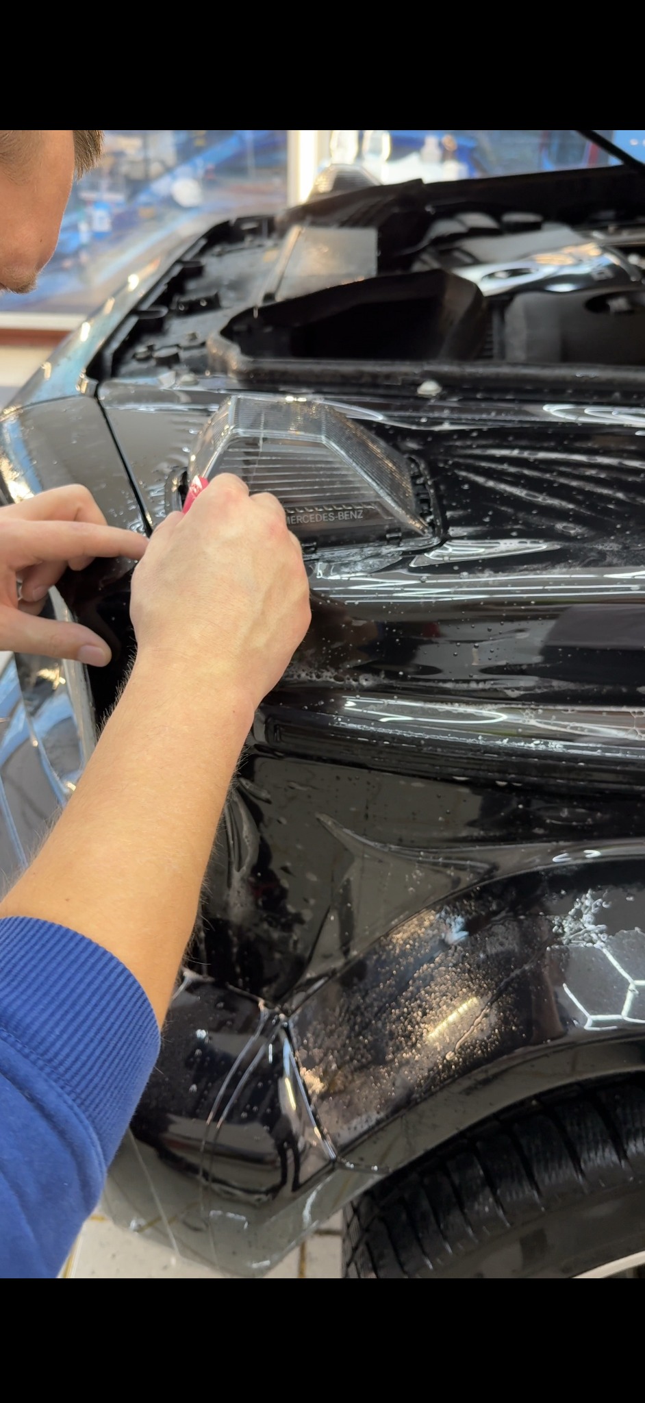 Nakładanie folii ochronnej na reflektor Mercedes-Benz. Widoczne dłonie aplikujące folię, w tle fragment karoserii. Ochrona reflektorów przed uszkodzeniami.