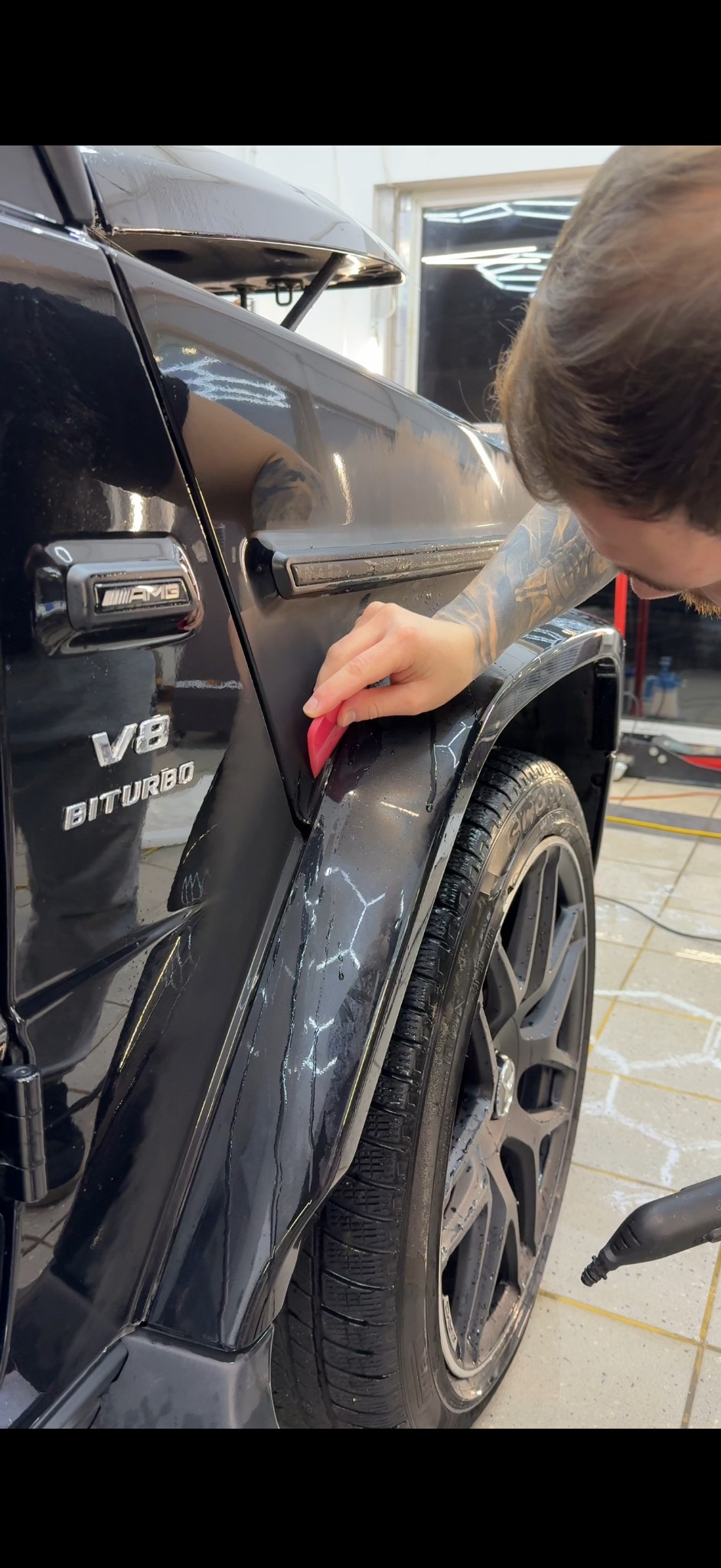 Czyszczenie błotnika czarnego Mercedesa G-klasy AMG V8 Biturbo w myjni. Widoczne ślady wody i piany, narzędzie w dłoni osoby z tatuażem na ramieniu.