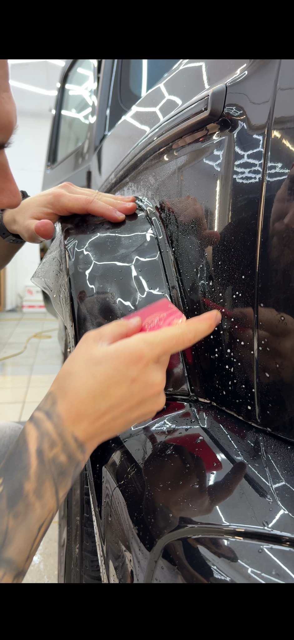 Oklejanie folią ochronną błotnika czarnego auta w Zielonej Górze. Widoczne dłonie aplikujące folię i rakla. Oświetlenie studyjne podkreśla detale.