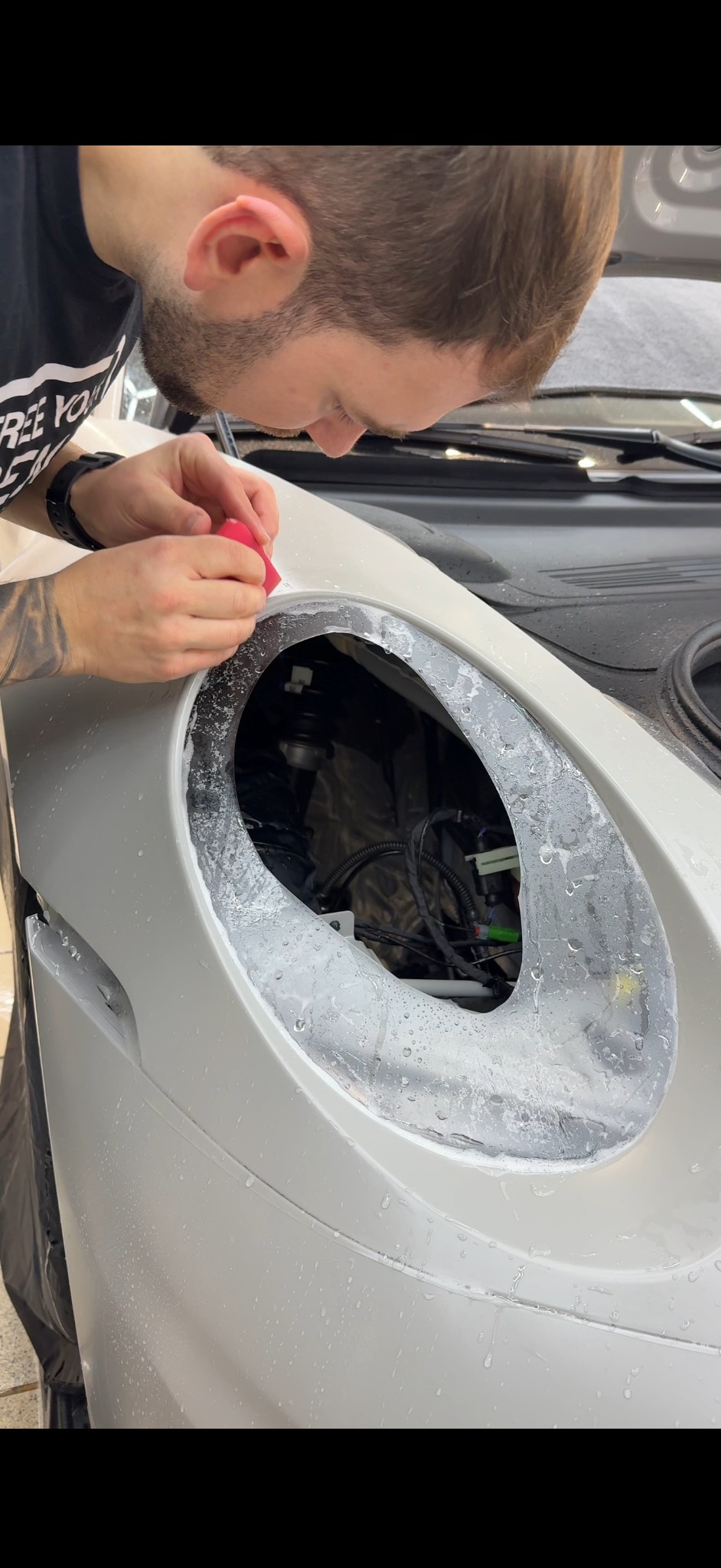 Aplikacja folii ochronnej na reflektor samochodu przez pracownika, widoczne narzędzia i woda na karoserii. Detailing auta w trakcie oklejania.