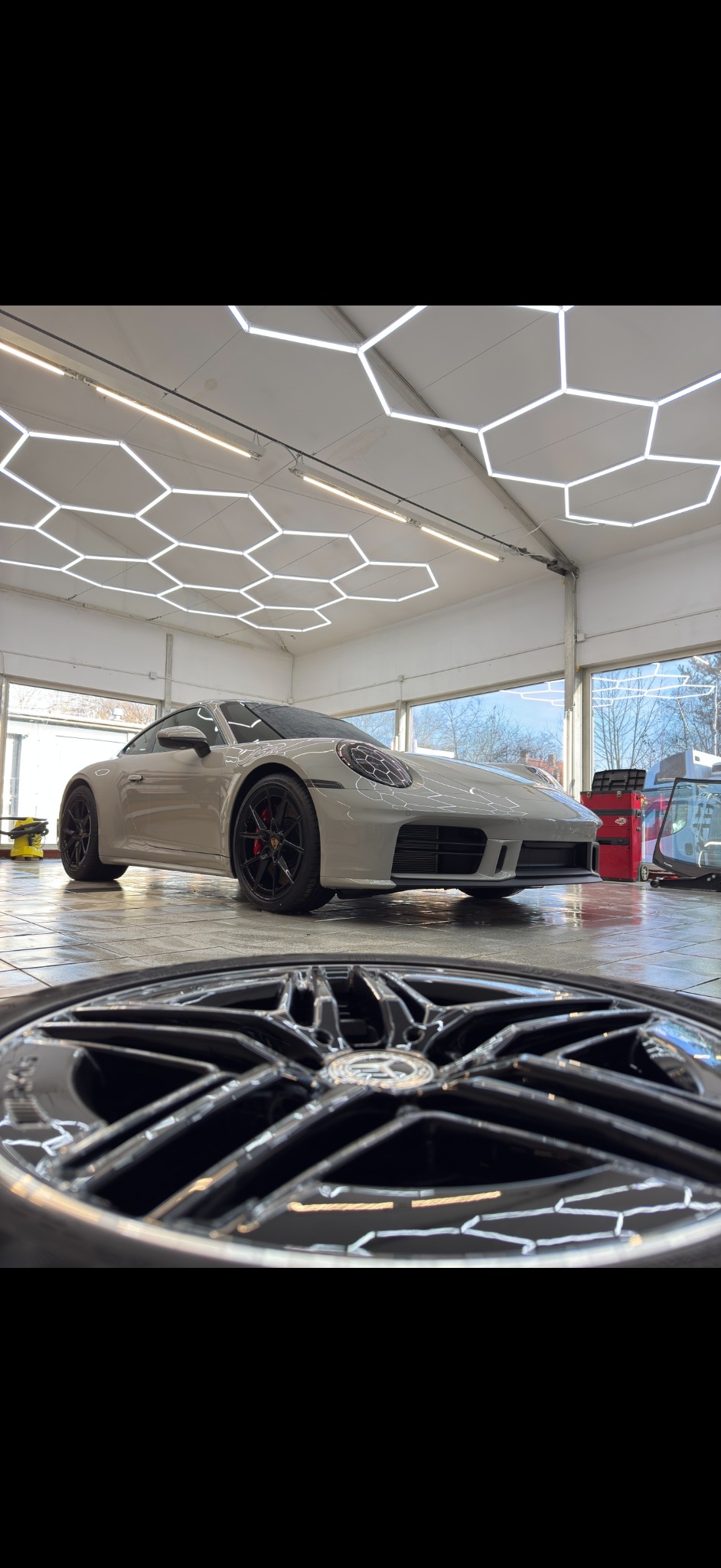 Białe Porsche 911 po detailingu, z czarnymi felgami na pierwszym planie, w jasnym pomieszczeniu z heksagonalnym oświetleniem sufitowym. Błyszcząca powierzchnia.