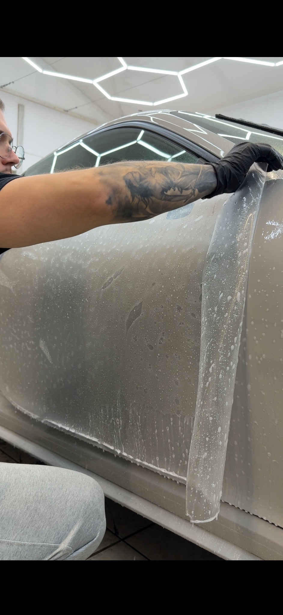 Nakładanie folii ochronnej na karoserię auta przez pracownika w rękawicach, widoczne krople wody i wzór oświetlenia sufitowego.