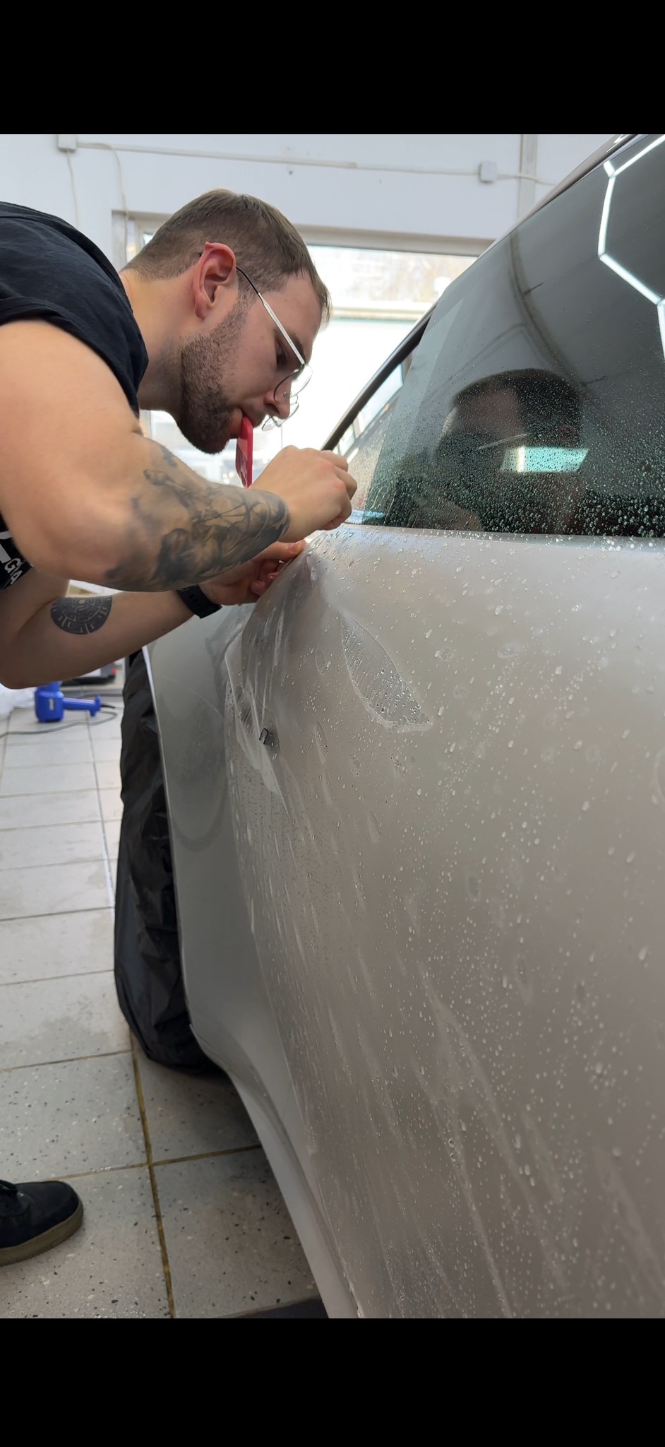 Pracownik w okularach aplikuje folię ochronną na karoserię jasnego auta, używając rakli. Widoczne krople wody i narzędzia. W tle oświetlenie warsztatowe.